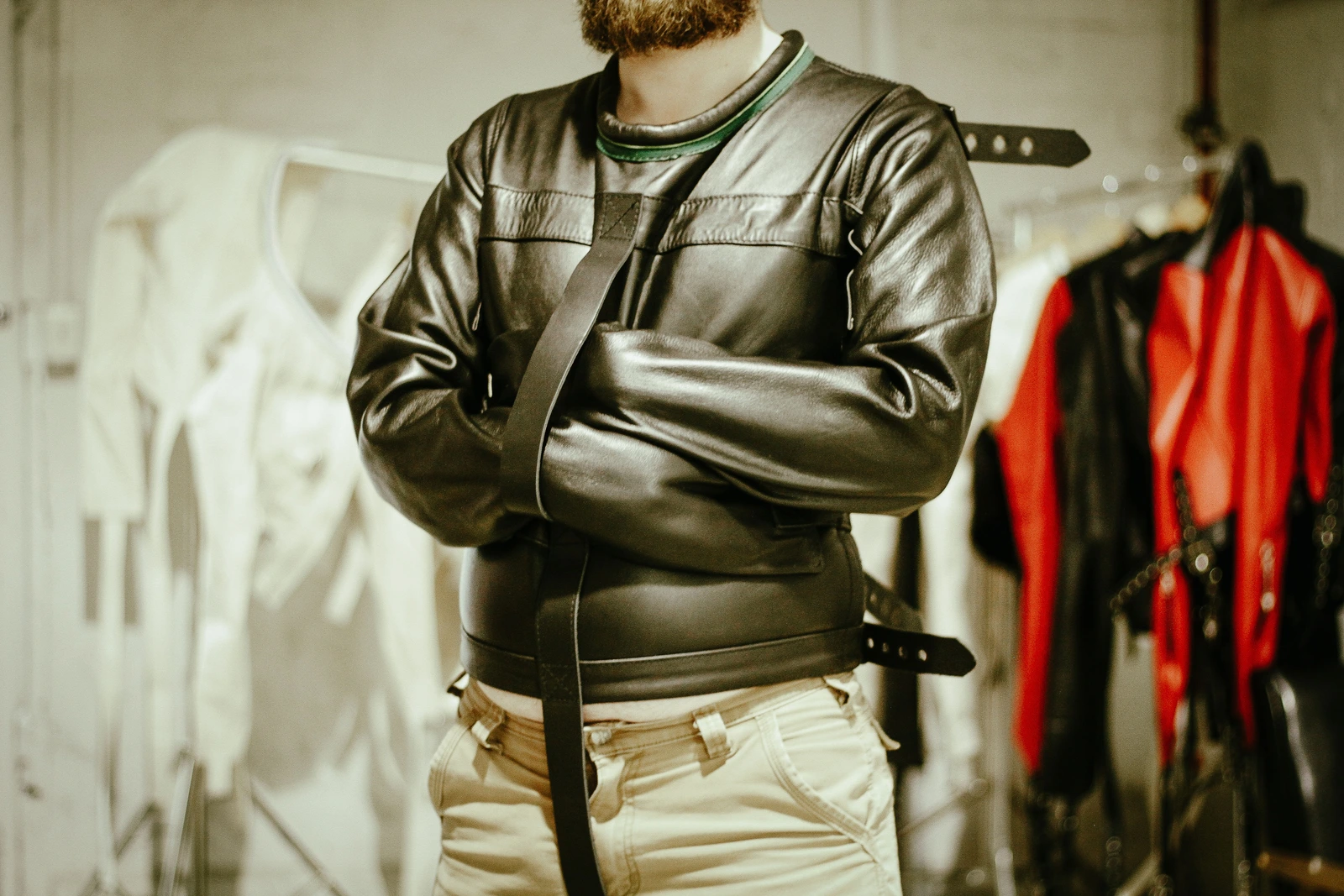 Leather Posey Replica with crossed sleeves photographed beside a rack of straitjackets.